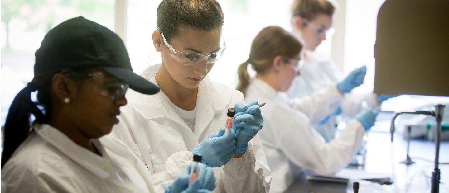 Students in white lab coats, safety glasses and blue gloves holding test tubes