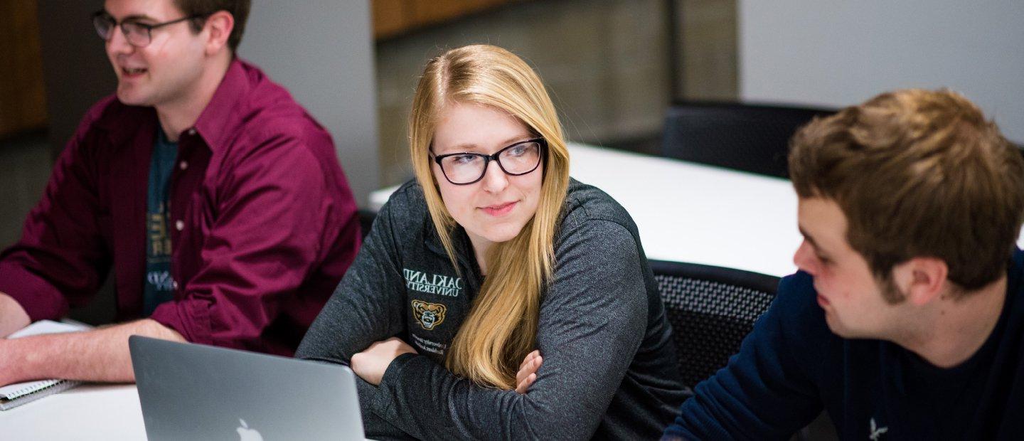 female student wearing a grey Oakland University shirt with an open laptop in front of her, male student on her right and left